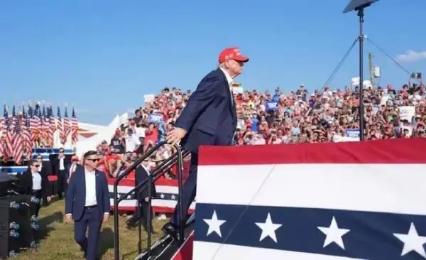 Trump is wearing a red hat and is preparing to walk up to the stage to give a speech, campaigning for votes in his presidential election.